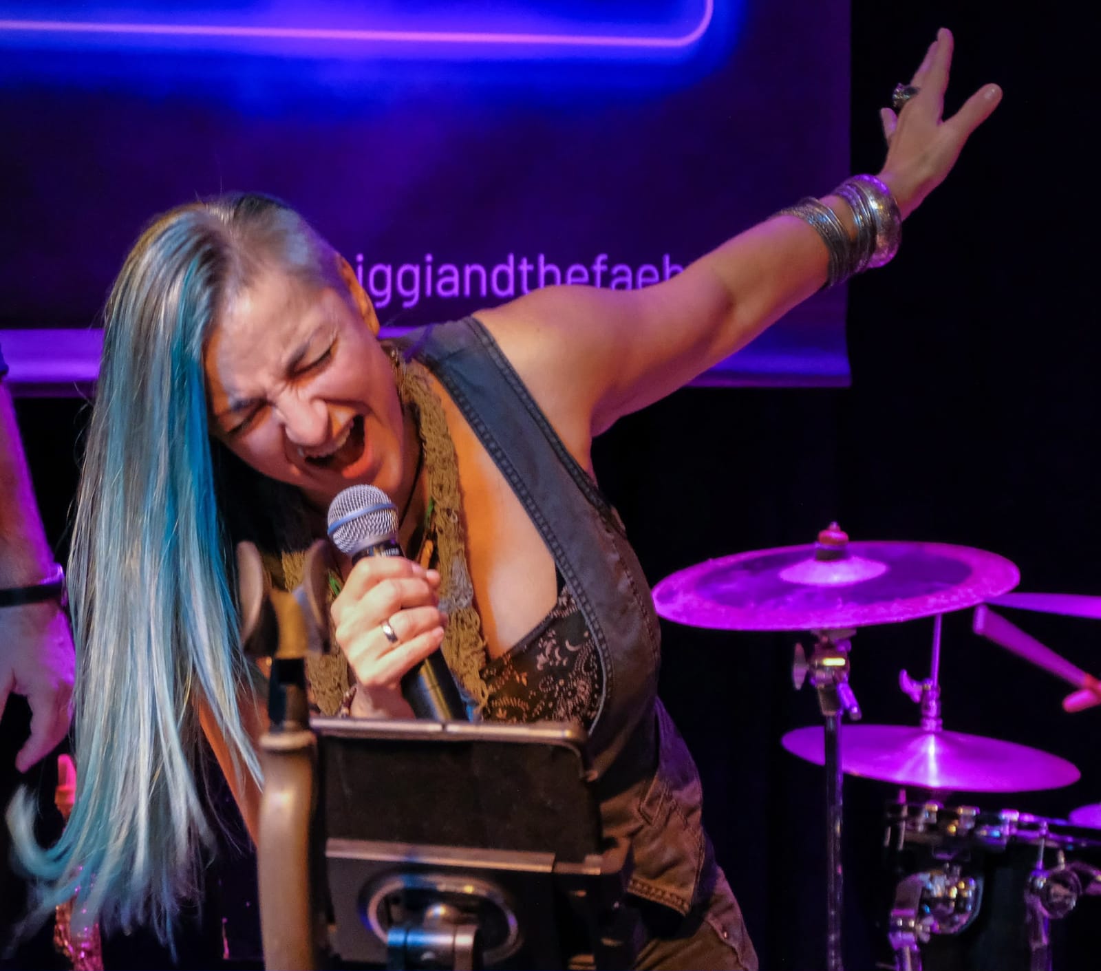 Biggi Käfer reißt singend den Arm hoch, hinter ihr das lila Neon-Banner von Biggi & the Fäbs und ein Schlagzeug
