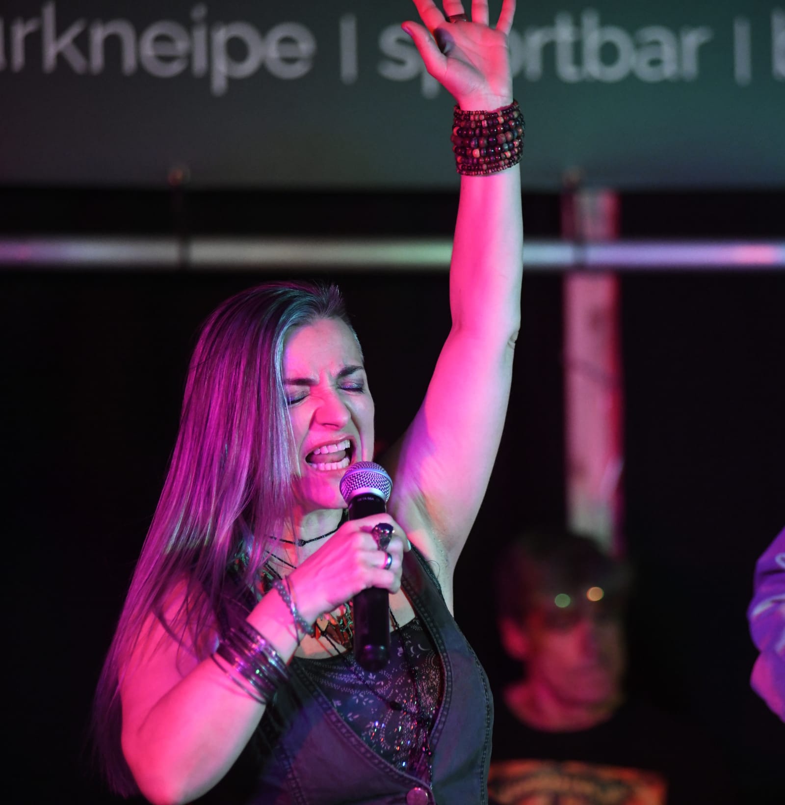 Biggi Käfer singt mit erhobenem Arm im magentafarbenen Bühnenlicht